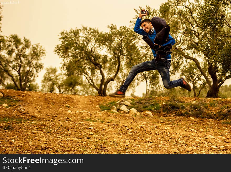 Man Wearing Blue and Black Shirt Jumping While Holding Brown Jacket Taken during Golden Hour