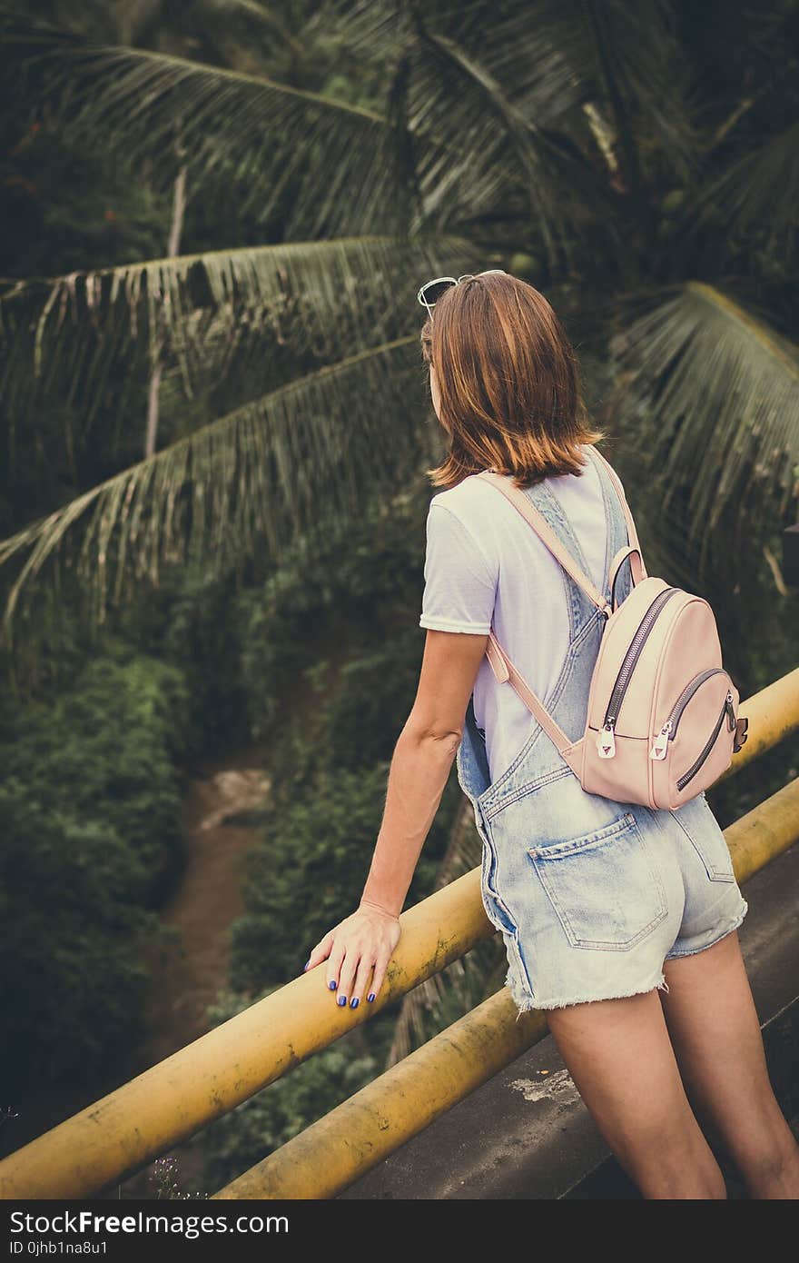 Woman Wearing Dungaree Shorts Stands Near a Yellow Metal Rail Overlook a River Belo With Coconut Trees at Daytime