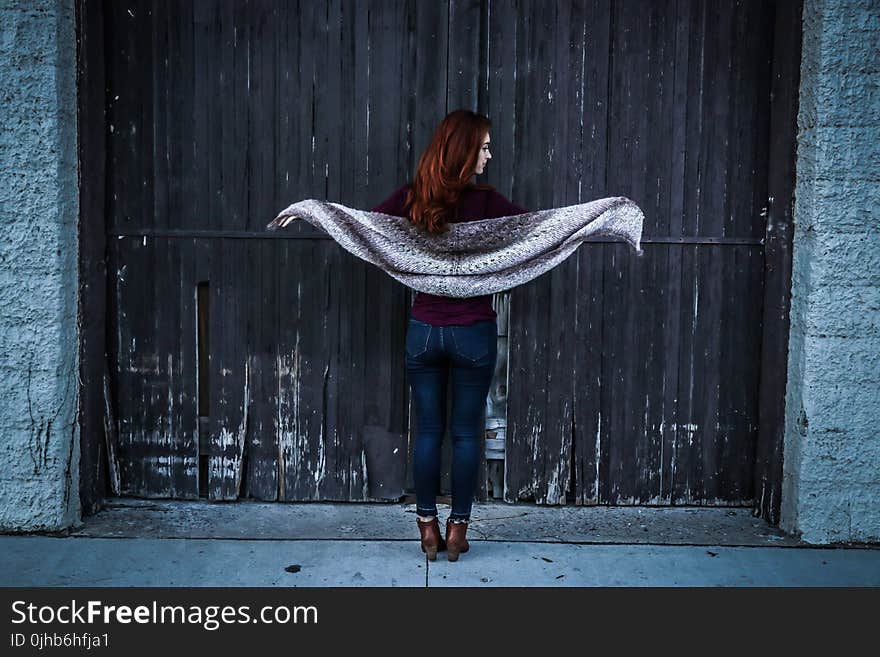 Woman Holding Gray Shawl While Spreading Her Arms Infront of Brown Wooden Door