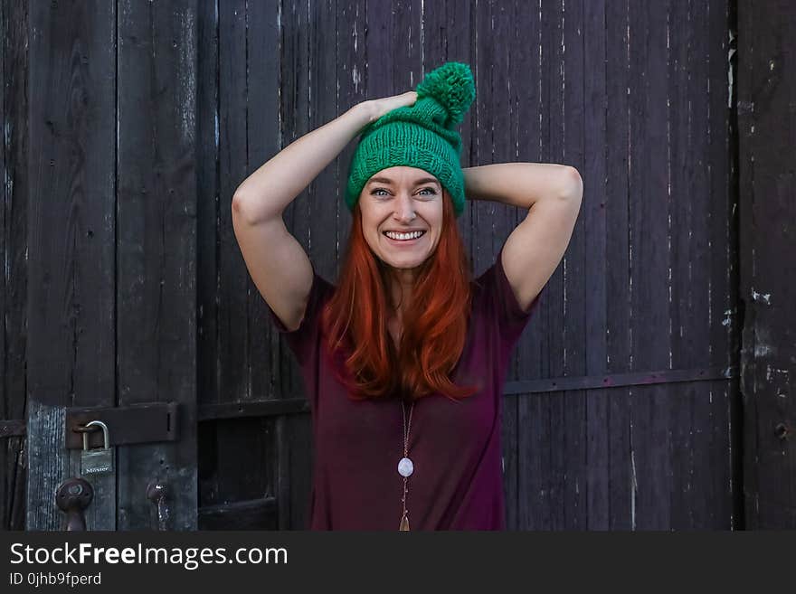 Woman in Green Bobble Cap and Purple T-shirt Posing Near Black Wooden Fence