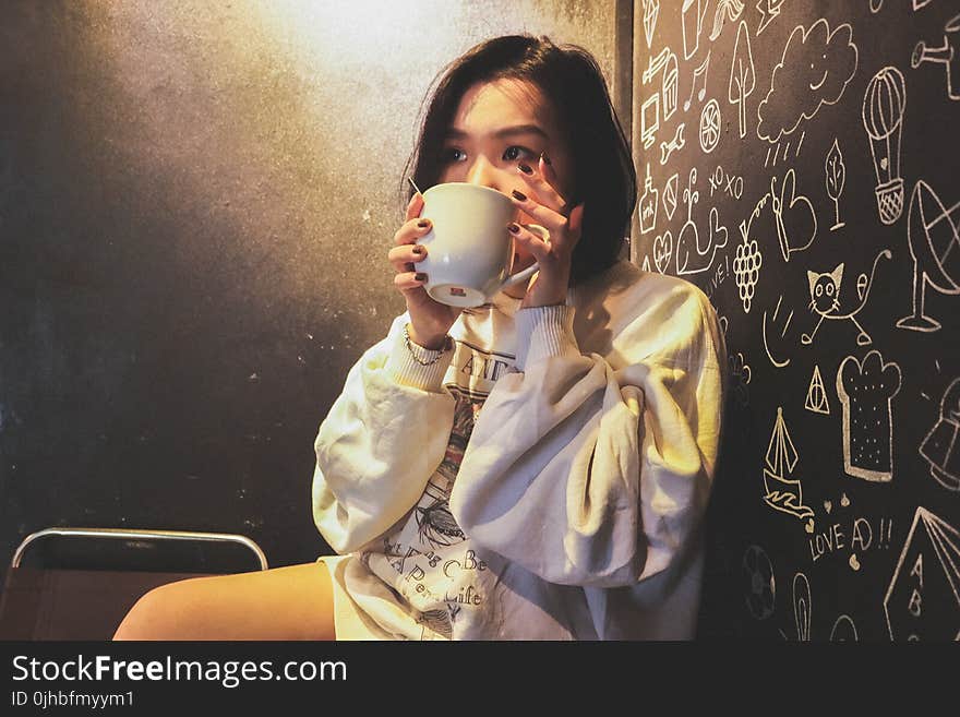 Woman Wearing White over Shirt Holding White Ceramic Mug Beside Black Printed Wall