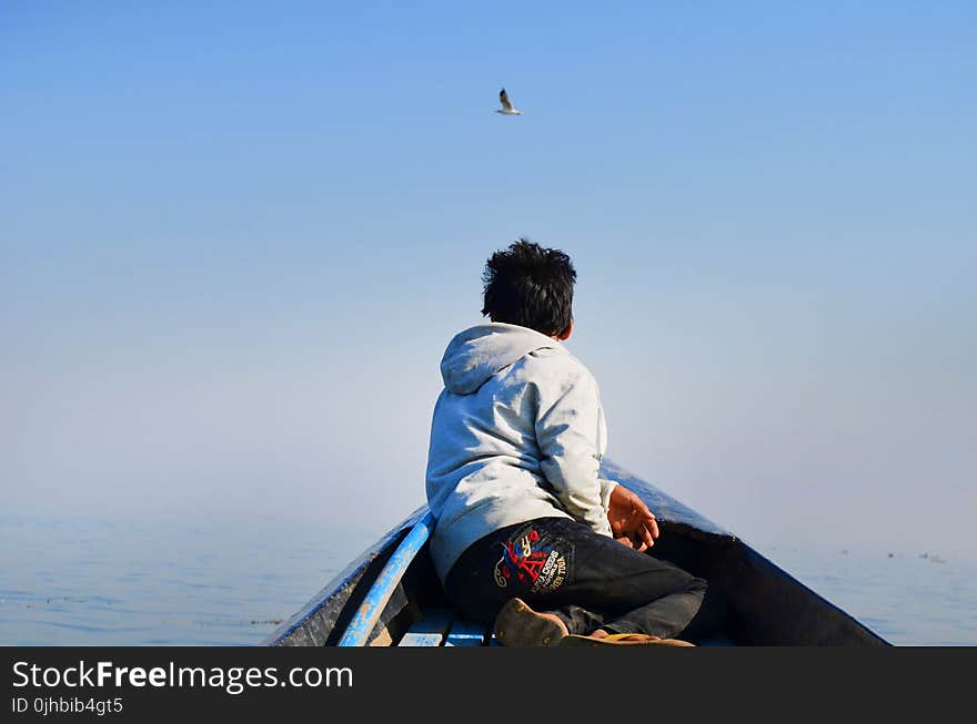 Man in Grey Hoodie and Black Pants Sitting in the Middle of Boat Looking at Bird in the Sky