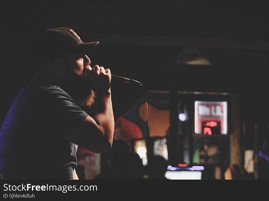 Man Wearing Grey Shirt and Black Cap Singing Using Corded Microphone