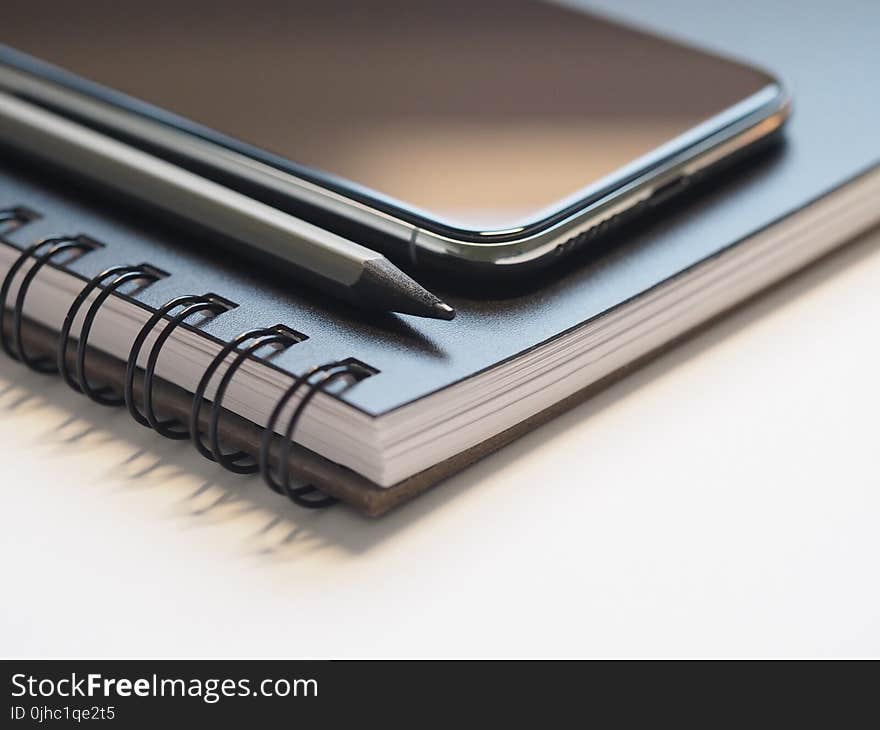 Closeup Photo of Black Smartphone Near Black and Grey Pencil on Black Spiral Notebook