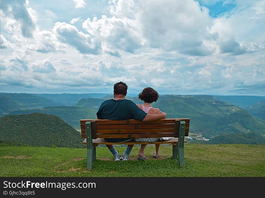 Couple Sitting on Brown Wooden Bench Near Mountains Covered With Grasses Under Blue Cloudy Sky