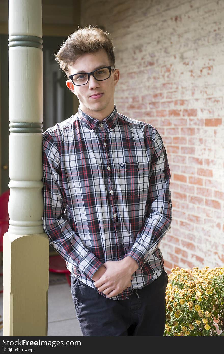 Blonde Man Wearing White, Gray, and Red Plaid Sport Shirt and Black Bottoms Standing While Leaning on Post