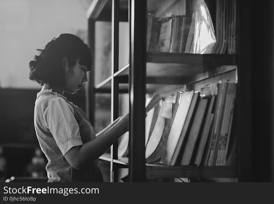 Grayscale Photo of a Woman Holding a Book Inside the Library