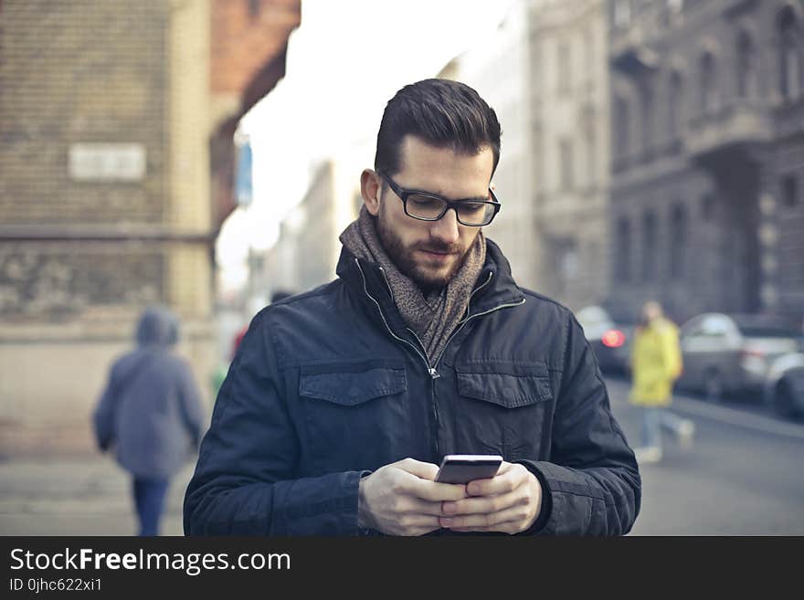 Man Wearing Black Zip Jacket Holding Smartphone Surrounded by Grey Concrete Buildings