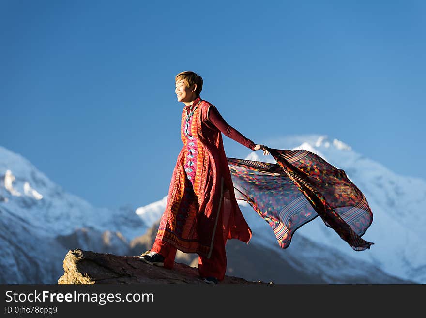 Woman in Orange Dress Standing on Top of Rock Cliff Holding Scarf Near Mountain Covered With Snow