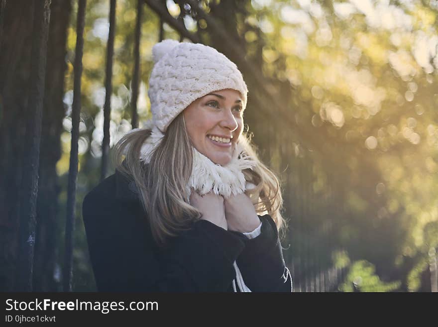 Photography of Woman in Black Jacket and White Knit Cap Smiling Next to Black Metal Fence