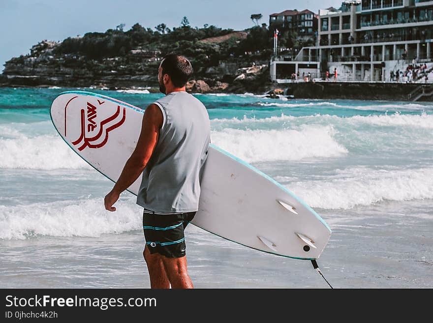 Man Walking Towards Body Of Water Holding A Surfboard