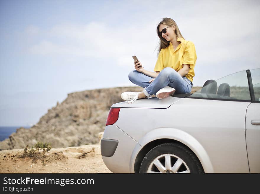 Woman in Yellow Blouse and Blue Jeans Taking Selfie While Sitting on Car