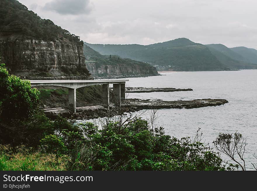 Concrete Bridge Near Mountain Above Shoreline Under Cloudy Sky