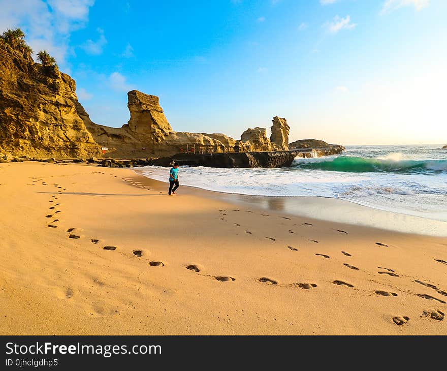 Person Walks on Brown Seashore Near Rock Formations