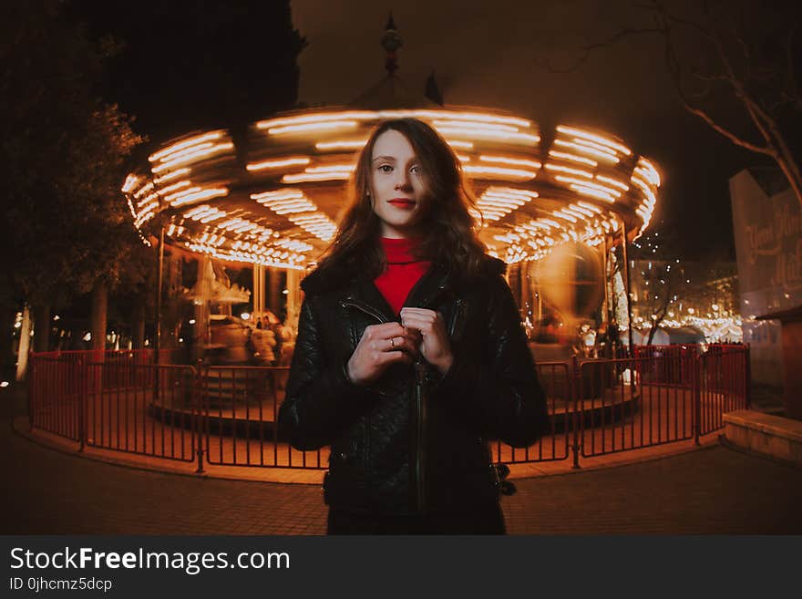 Woman Wearing Black Leather Jacket and Red Turtle-neck Shirt Standing in Front of Carousel Ride