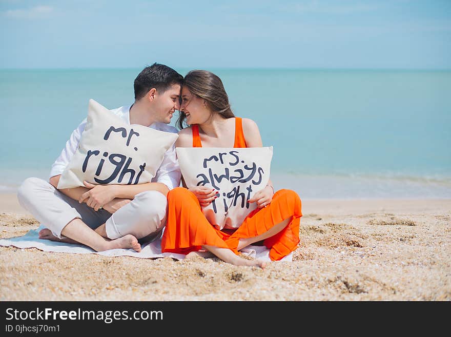 Man and Woman Wearing Cloths Sitting on Brown Sand Near Seashore