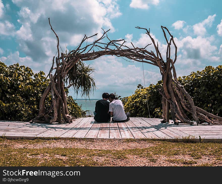 Man and Woman Sitting on Wooden Dock Under Cloudy Sky