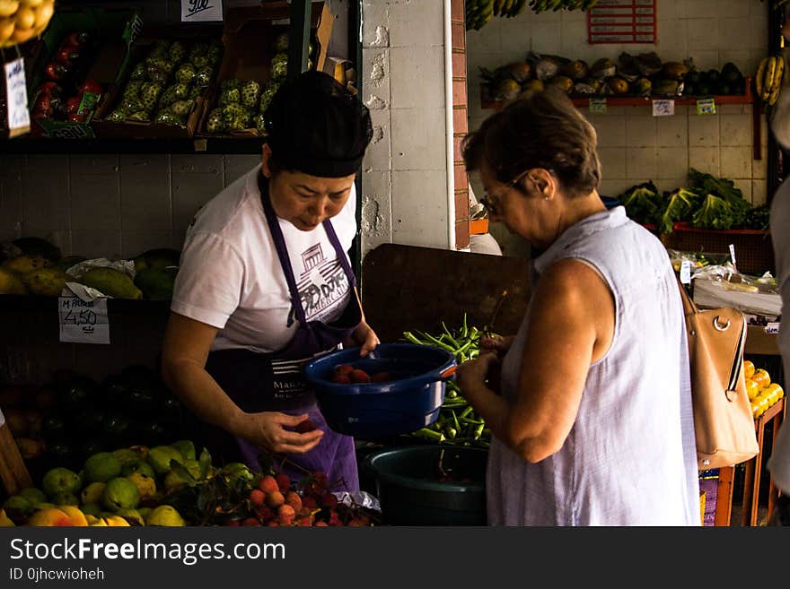 Photo of Woman Holding Blue Bucket and Fruit Facing Woman Wearing Blue Sleeveless Shirt