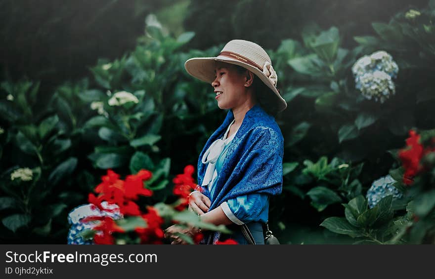 Selective Focus Photo of Woman in Blue Shawl and Brown Sun Hat in the Middle of Garden