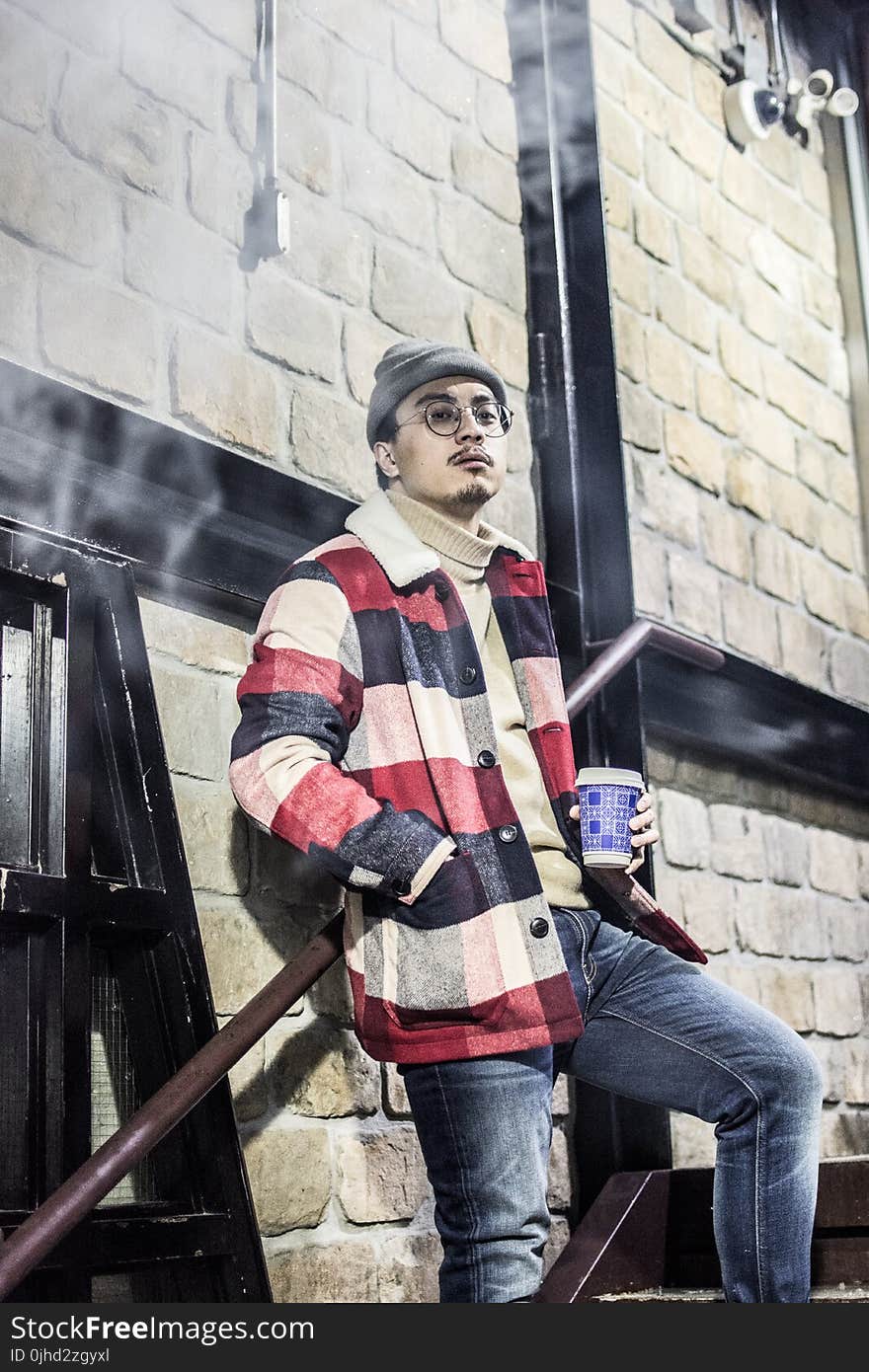 Man in Red, Black, and Grey Plaid Button-up Jacket With Blue Denim Jeans Outfit Holding Disposable Cup Stands Beside Brown Concrete Wall