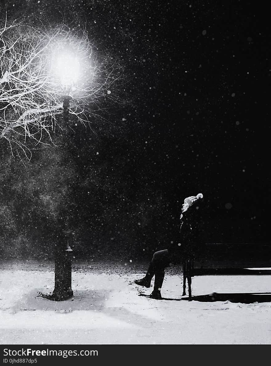 Grayscale Photography of Person With Knit Pompom Cap Sits in Front Turned on Light Post at Night