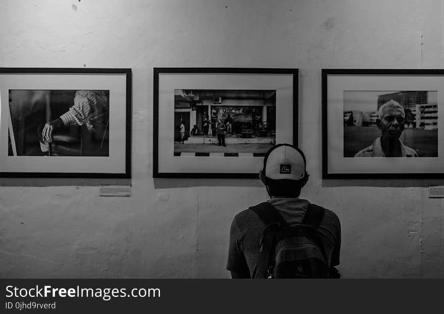 Grayscale Photo of Man Wearing White Cap in Front of Three Paintings