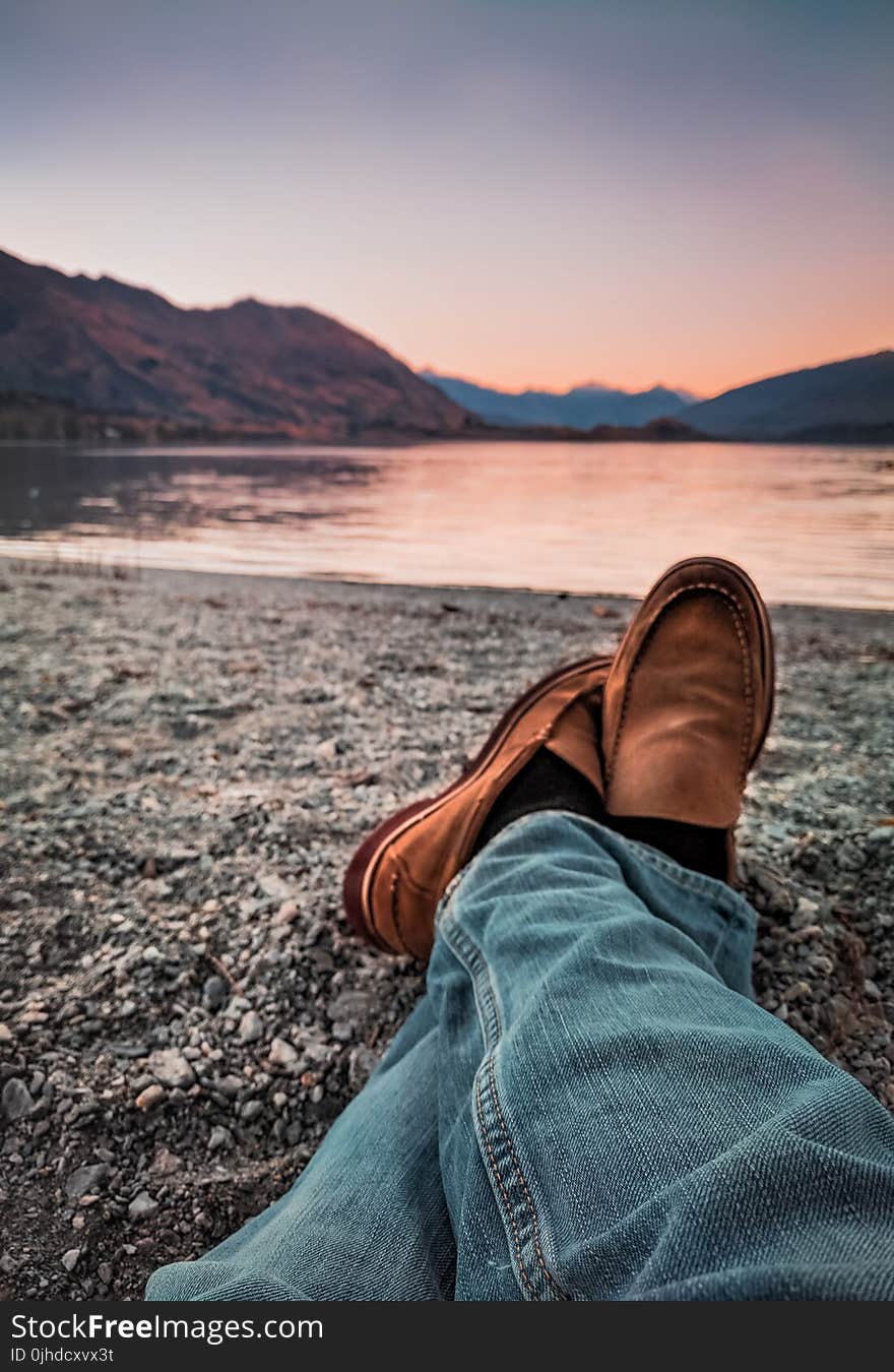 Person Wearing Blue Jeans and Brown Leather Loafers Sitting Beside Gray Sand