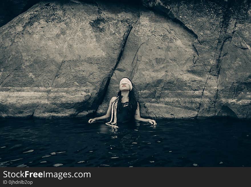 Woman in Blindfold Wearing Black Top on Body of Water While Leaning on a Rock