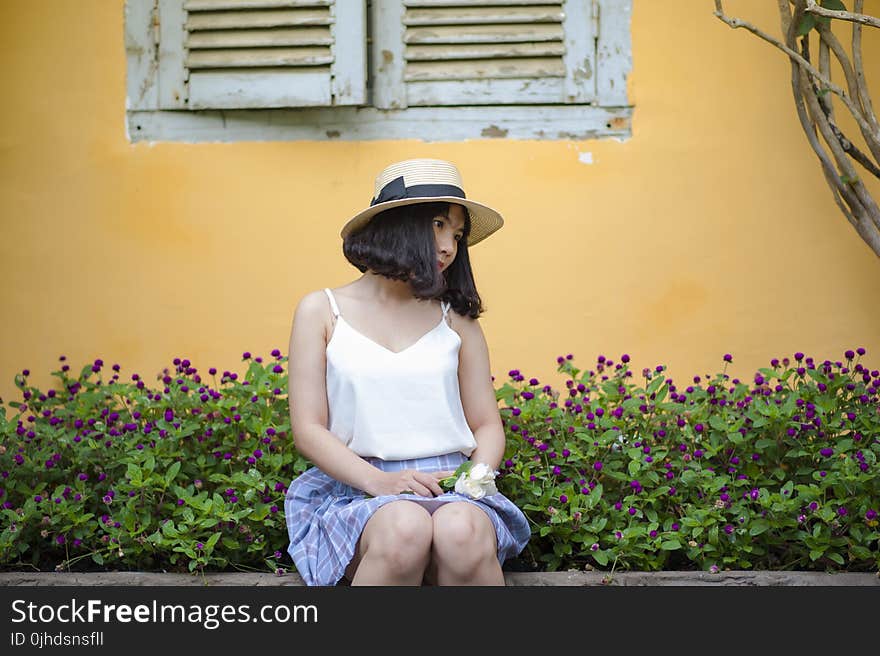 Woman in White Spaghetti Strapped Top With Blue and Purple Plaid Skirt Sitting on Ledge Near Purple Petaled Flower Garden Posing for Photo