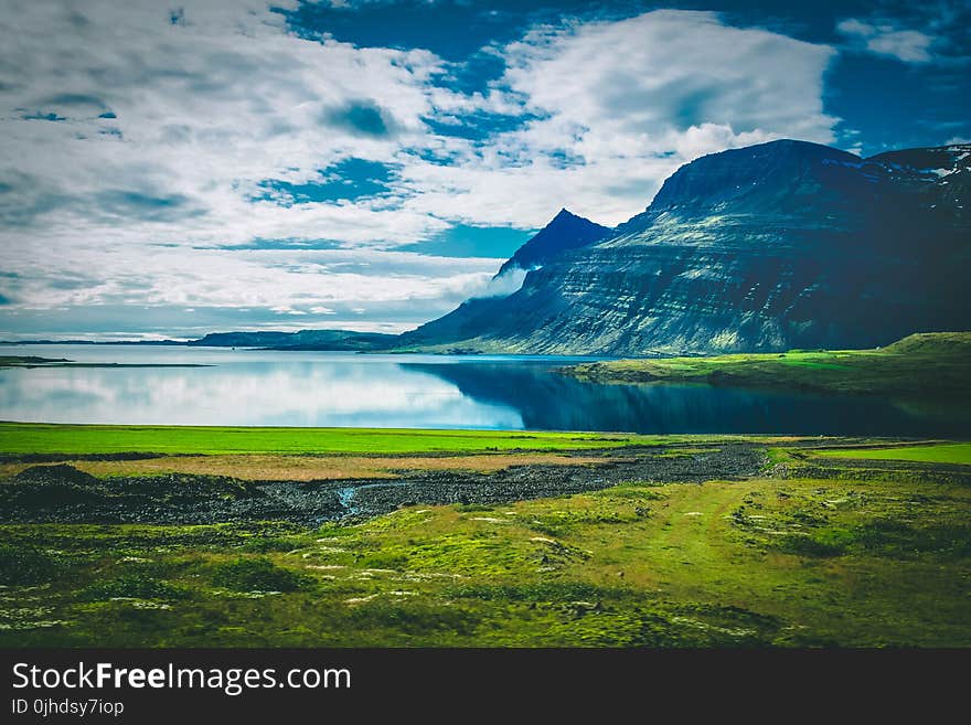 Calm Body of Water Near Black Mountain Surrounded by Green Grass