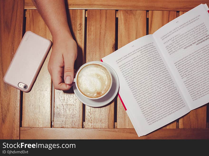 Person&#x27;s Hand Holding White Coffee Mug With Plate on Brown Wooden Board With White and Black Book