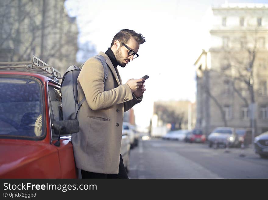 Man in Beige Coat Holding Phone Leaning on Red Vehicle