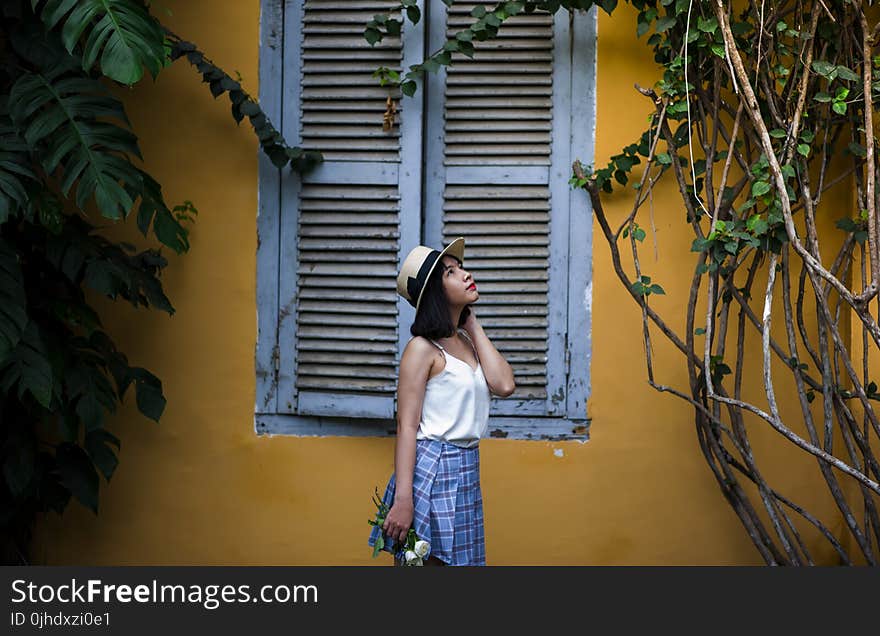 Photo of Woman Wearing White Sleeveless Top and Hat Holding Flowers Near Window