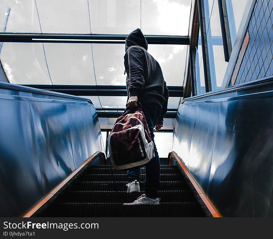 Person Wearing Black Hooded Jacket Standing on Escalator While Holding Backpack