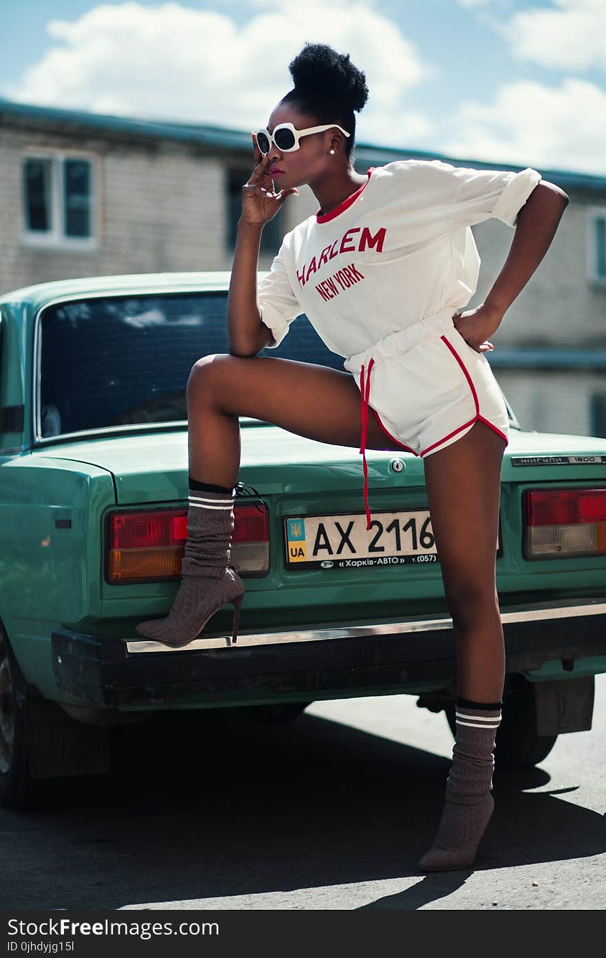 Photo of Woman Wearing White Long-sleeved Shirt and White Dolphin Shorts With Red Trim Standing Near Teal Car