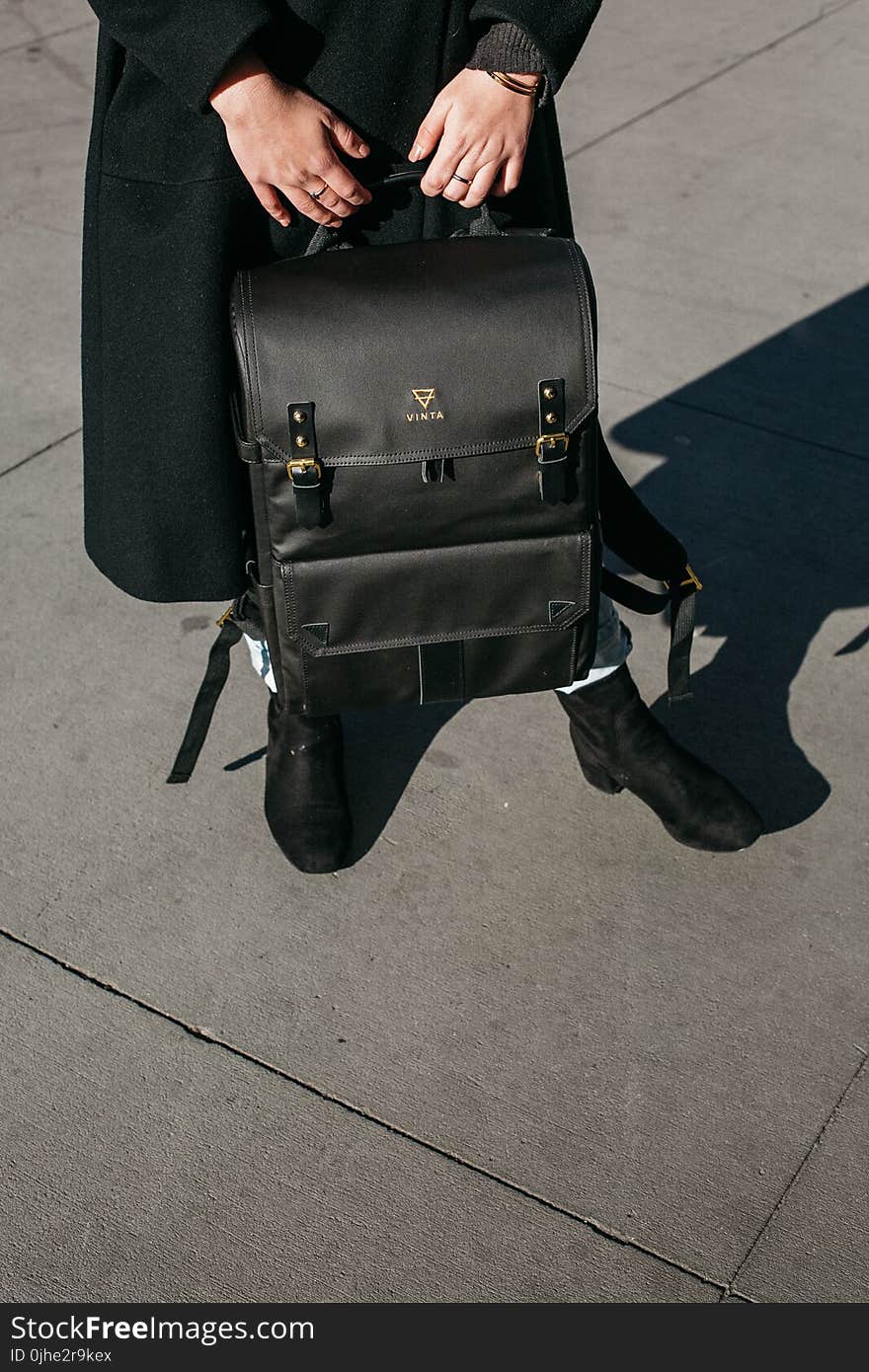 Person Wearing Black Coat Holing Black Leather Knapsack Backpack Standing on Concrete Ground