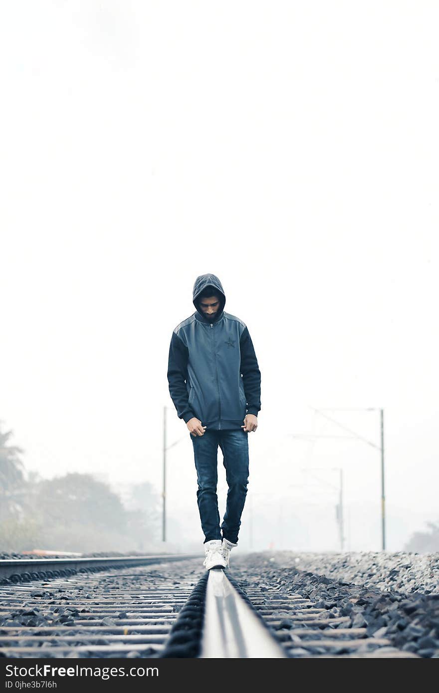 Man Wearing Gray and Black Zip-up Hoodie With Black Denim Jeans and White Shoes Walking on Train Railing Behind White Fog