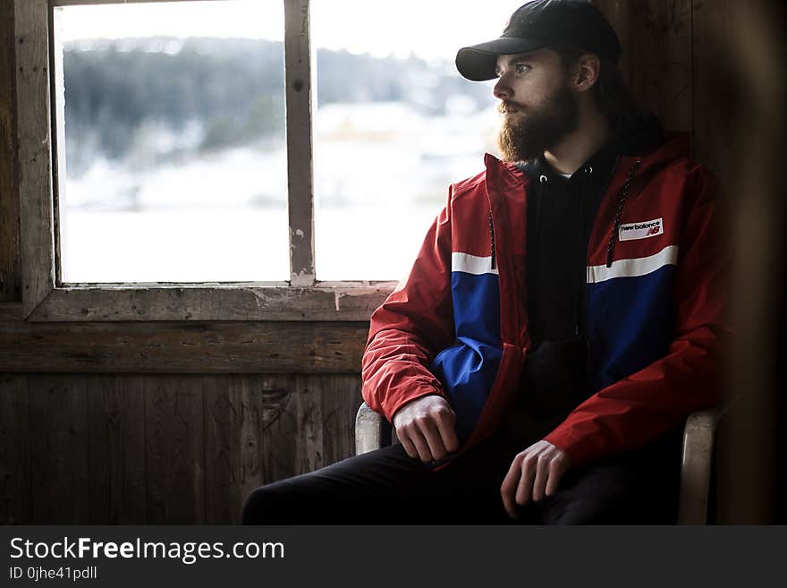 Photo of Man Wearing Black Hat and Blue-and-red Jacket Near Window