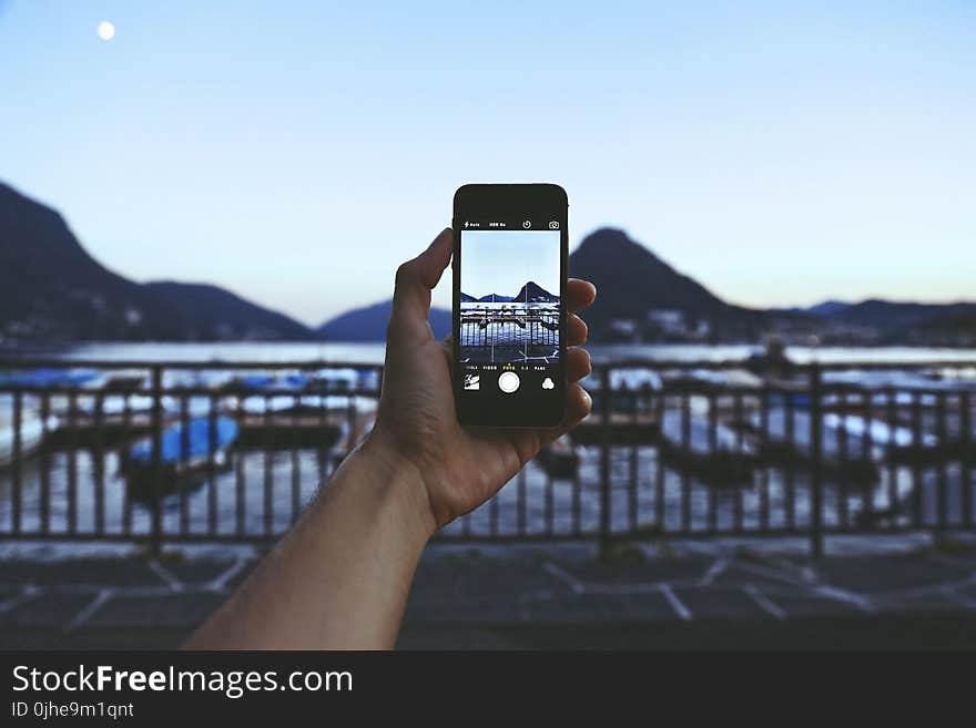 Person Holding Iphone Taking Picture of Mountain Near Body of Water