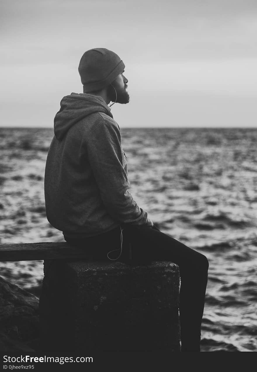 Grayscale Photo of Man in Hoodie and Kit Cap Sitting Near Bodies of Water