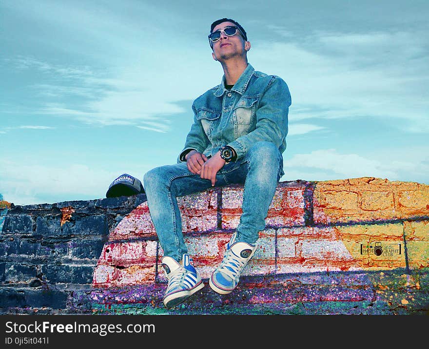 Man in Gray Denim Jacket and Jeans Sitting on Brick Wall