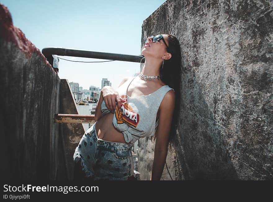 Woman Wearing White and Orange Tank Top and White Denim Shorts Leaning of Gray Concrete Pavement