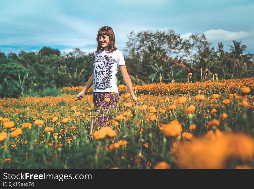 Woman in White and Black Floral Crew-neck T-shirt and Red Bottoms Standing on Orange Petaled Flower Field at Daytime