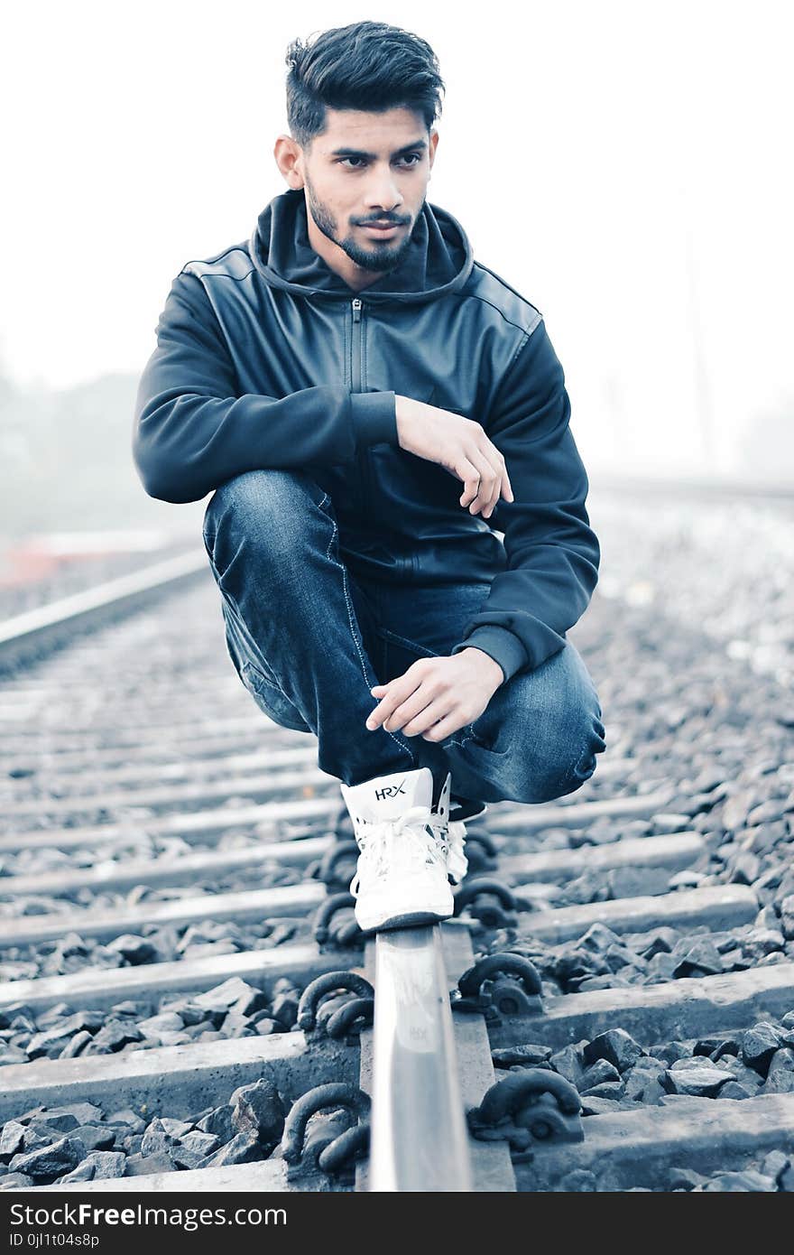 Man in Black Leather Zip-up Hoodie and Black Denim Jeans Sitting on Grey Metal Train Railings Surrounded With Rocks during Husky Morning