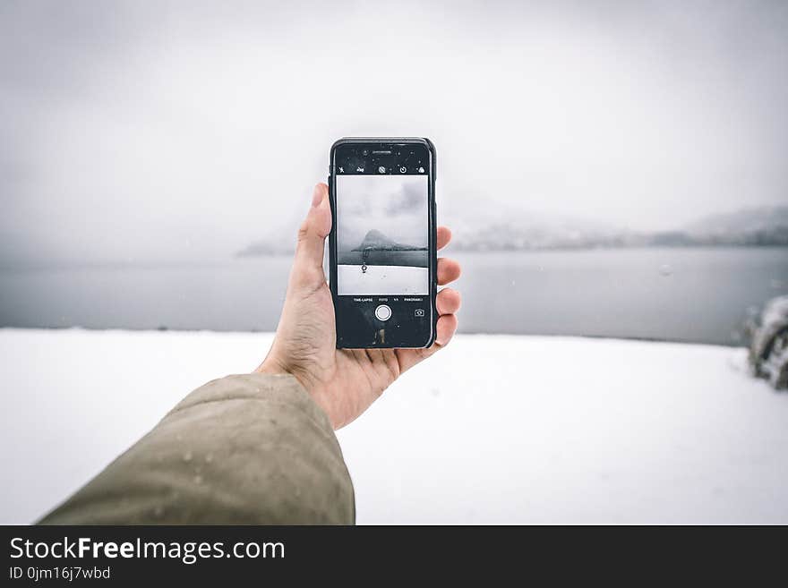 Photo of Person Wearing Brown Coat Holding Android Smartphone While Taking Picture of Mountain and Body of Water