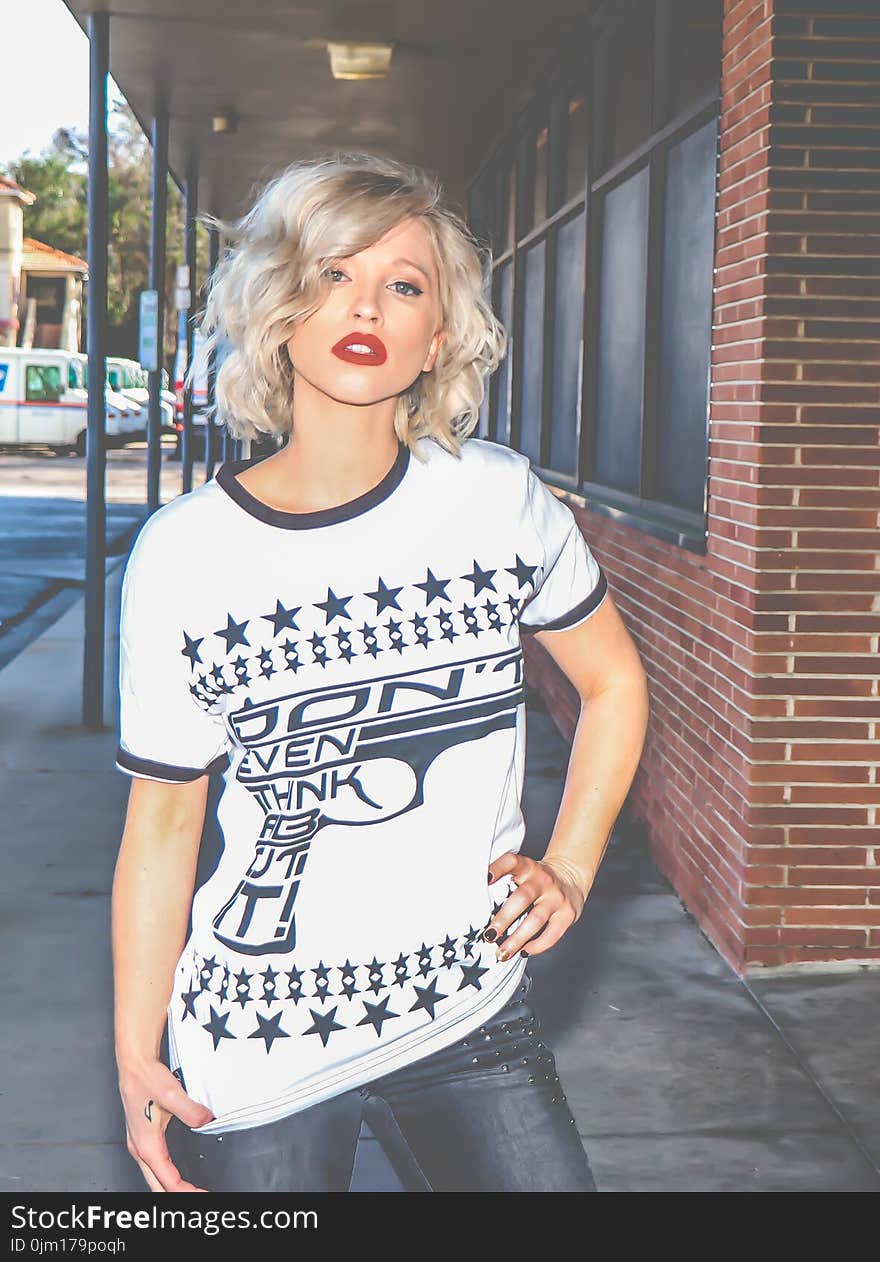 Woman in White and Black Crew-neck T-shirt Stands on Pathway Near Red Bricked Building