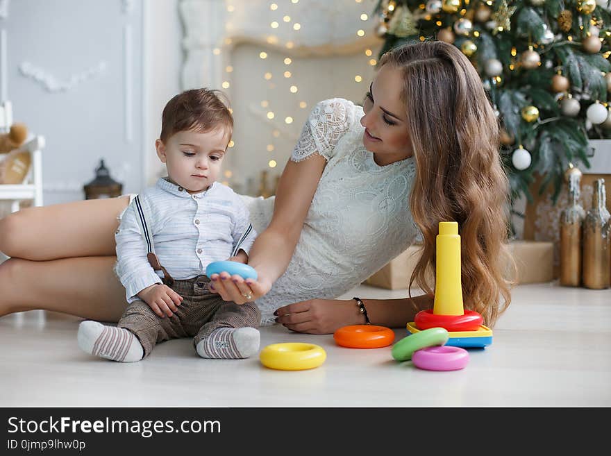 Young women with long hair,dressed in a short white lace dress lies on white floor with sitting next to a young son,dressed in a white shirt and grey trousers,mother teaches son how to assemble a pyramid,the back is green decorated Christmas tree