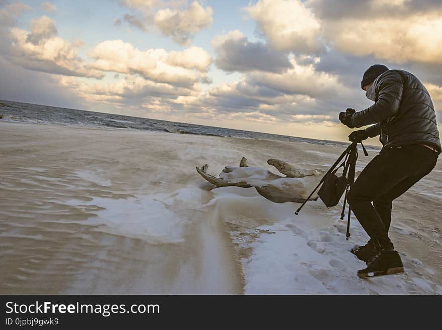 Man in Black Bubble Jacket Walking on Seashore While Holding Black Camera Tripod