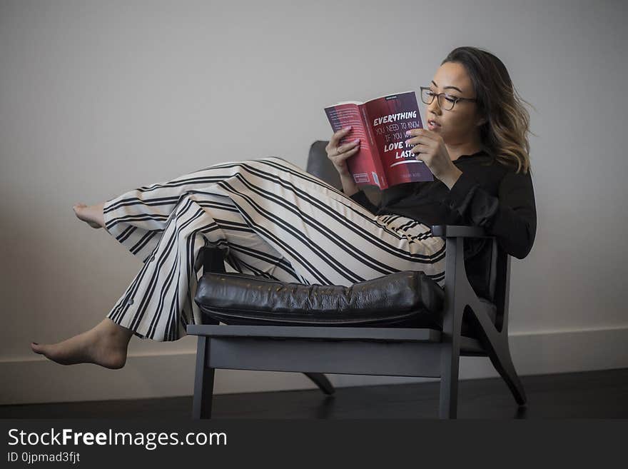 Women&#x27;s Black Long-sleeved Top, White and Black Striped Pants Reading Book Sitting on Gray Wooden Framed Padded Armchair