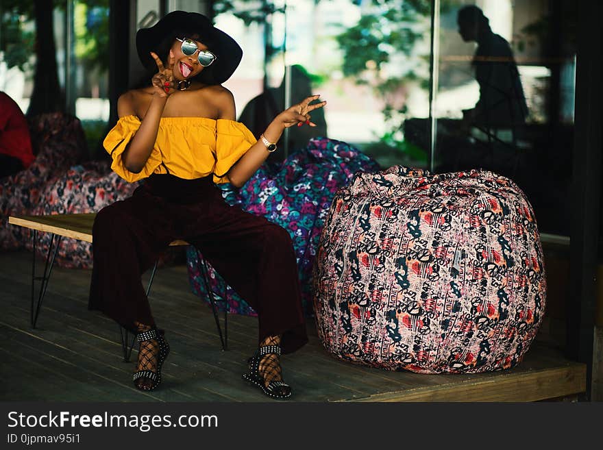 Woman Yellow Off-shoulder Top and Black Pants Sitting on Brown Bench With Tongue Out